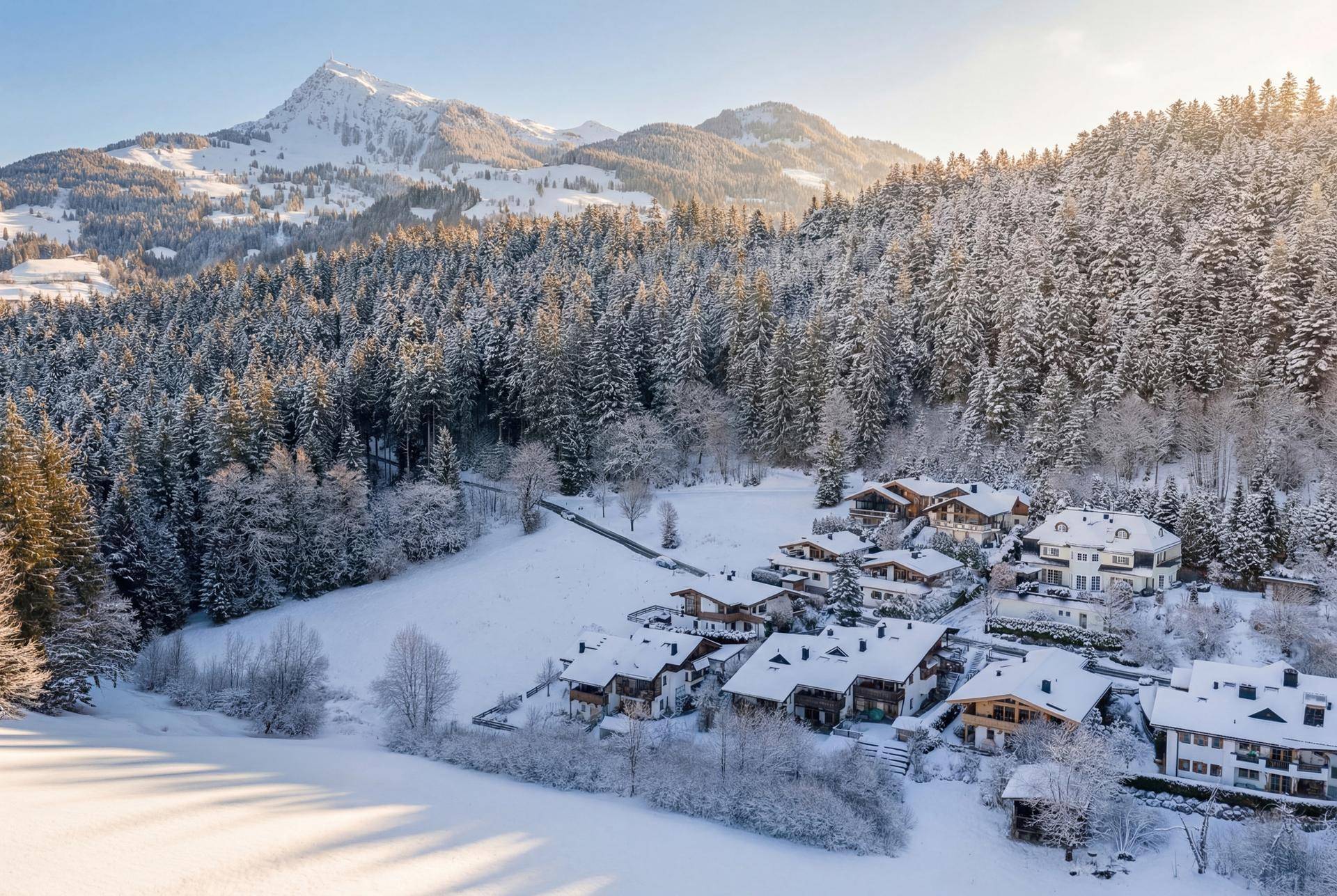 Panoramablick auf eine verschneite Siedlung am Waldrand mit majestätischen Bergen im Hintergrund.
