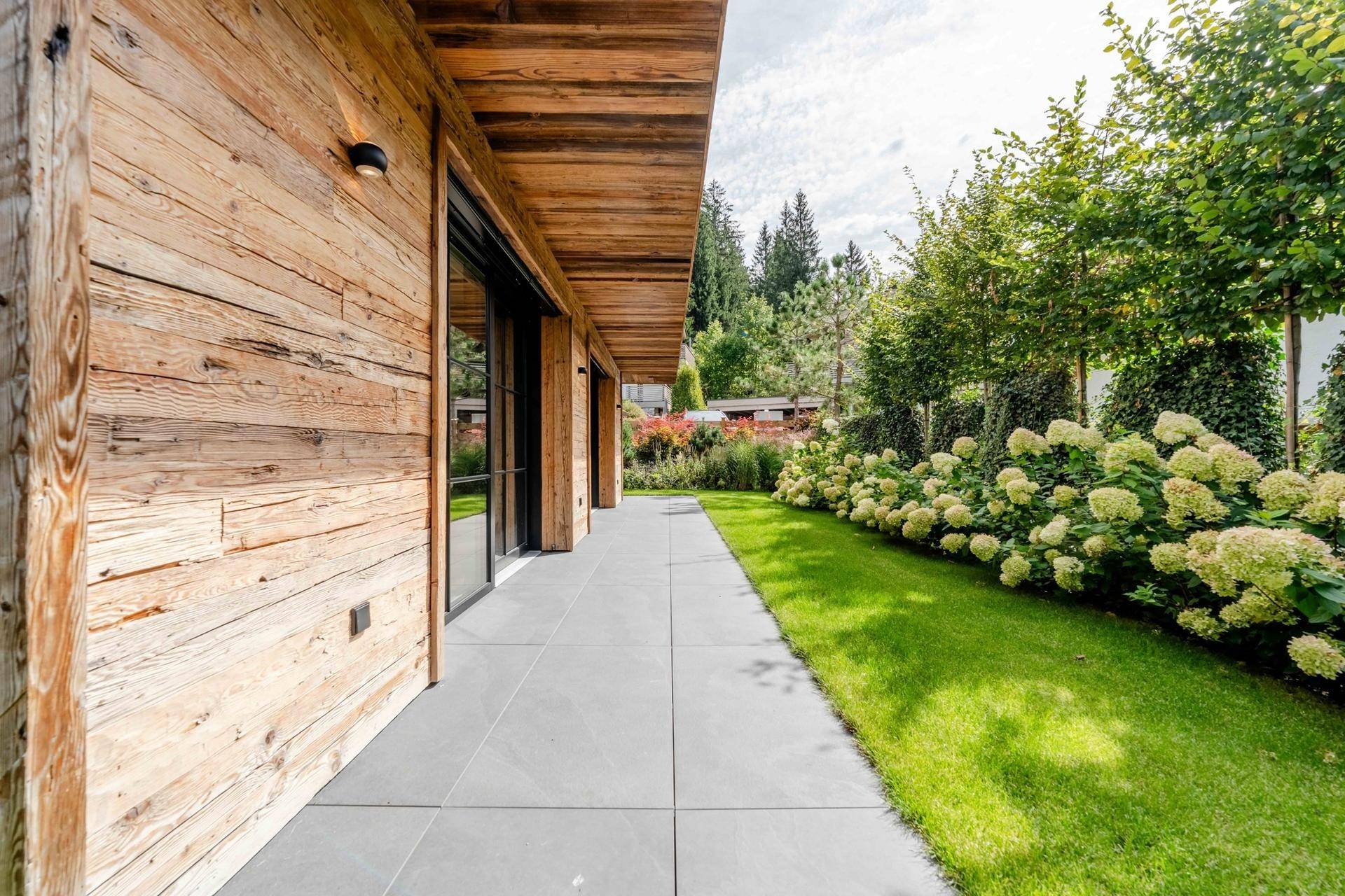 Gepflegter Gartenbereich mit Rasenfläche, blühenden Sträuchern und einer modernen Holzfassade.