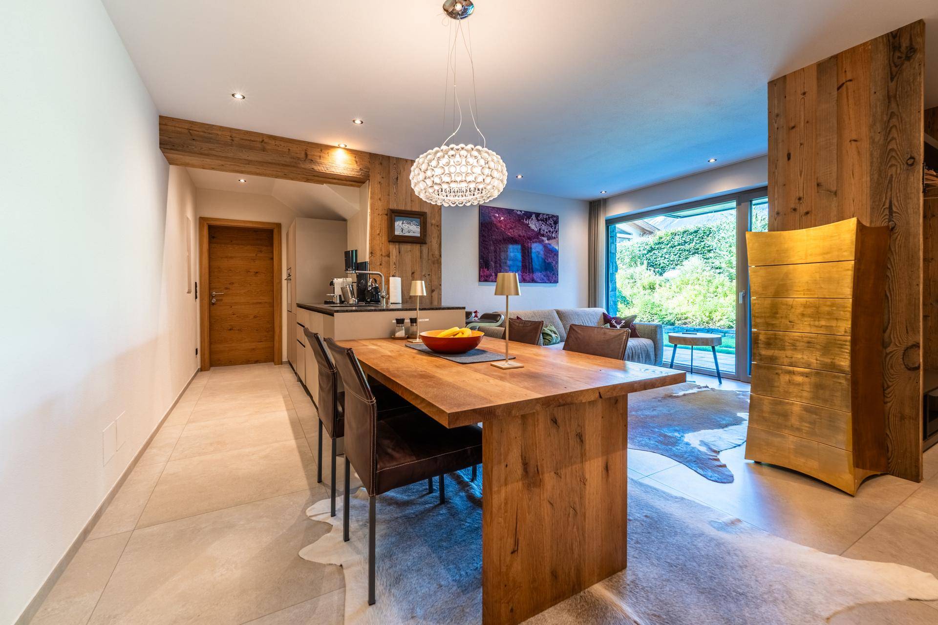 Rustikaler Essbereich mit Holztisch, Kuhfellteppich und kleiner Küchenzeile im Hintergrund.