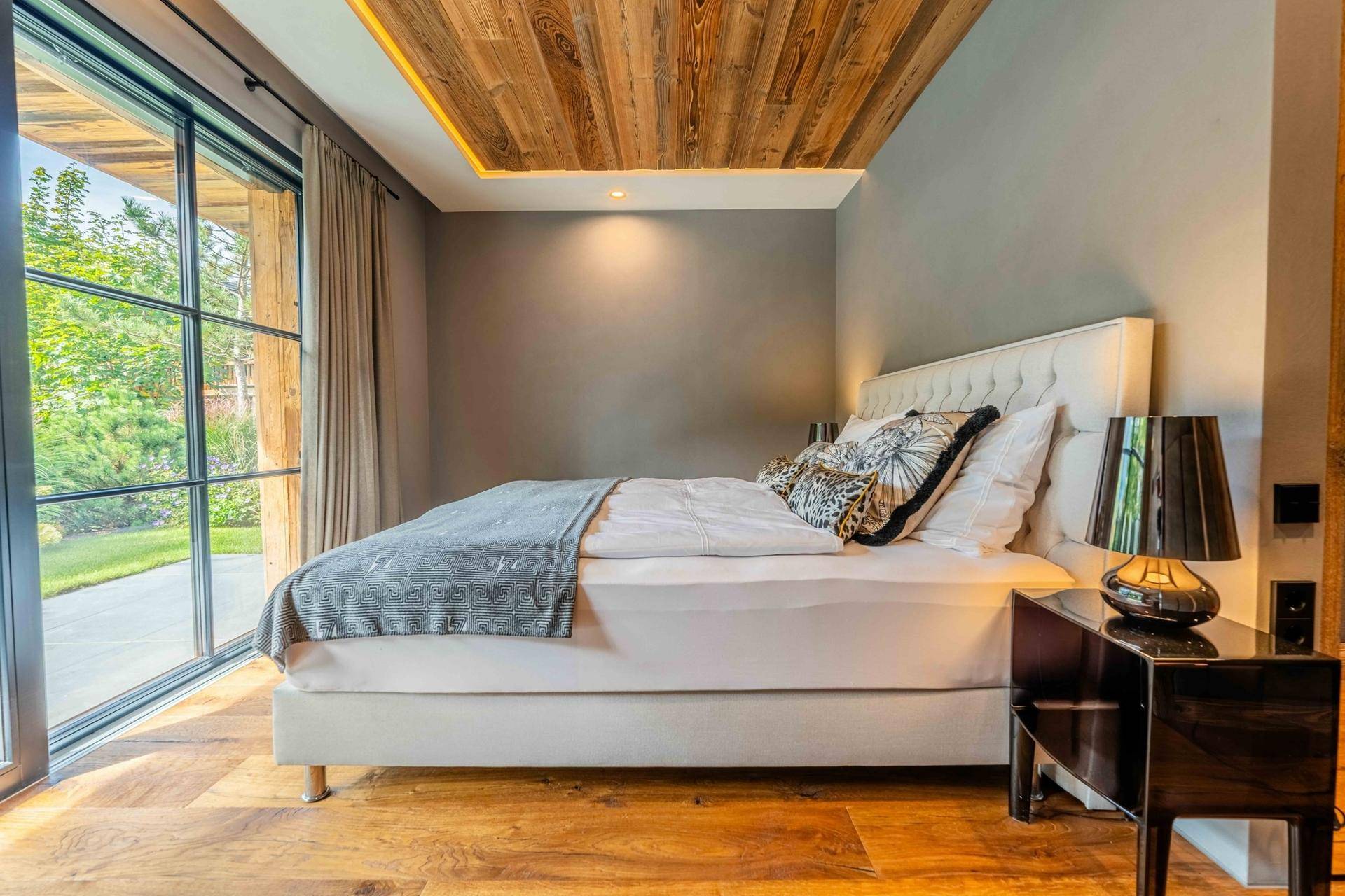 Helles Schlafzimmer mit Holzboden, Holzdecke und großen Fenstern mit Blick ins Grüne.