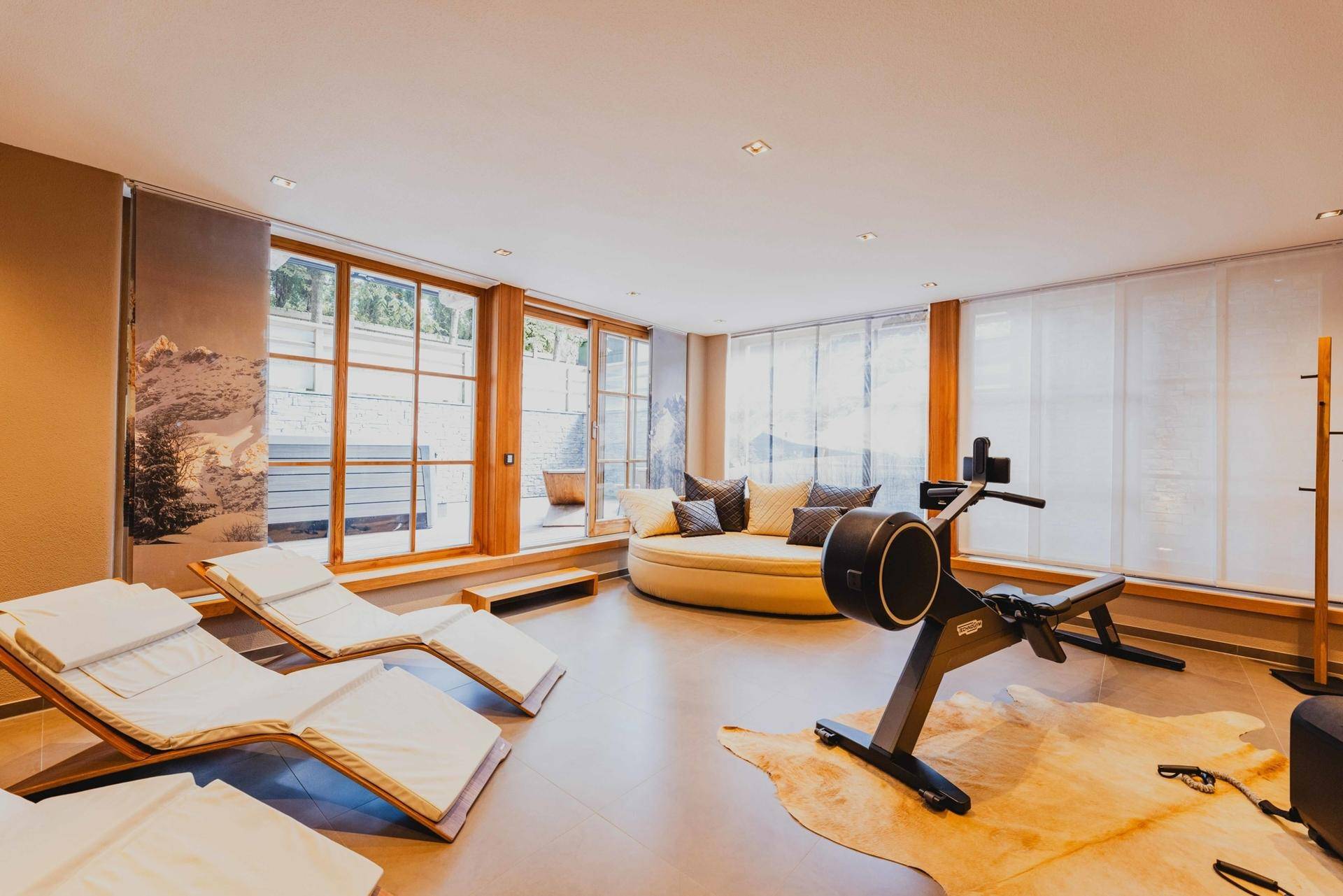 Heller Fitnessraum mit Liegeflächen, Rudergerät und großen Fenstern, die den Blick nach draußen freigeben.