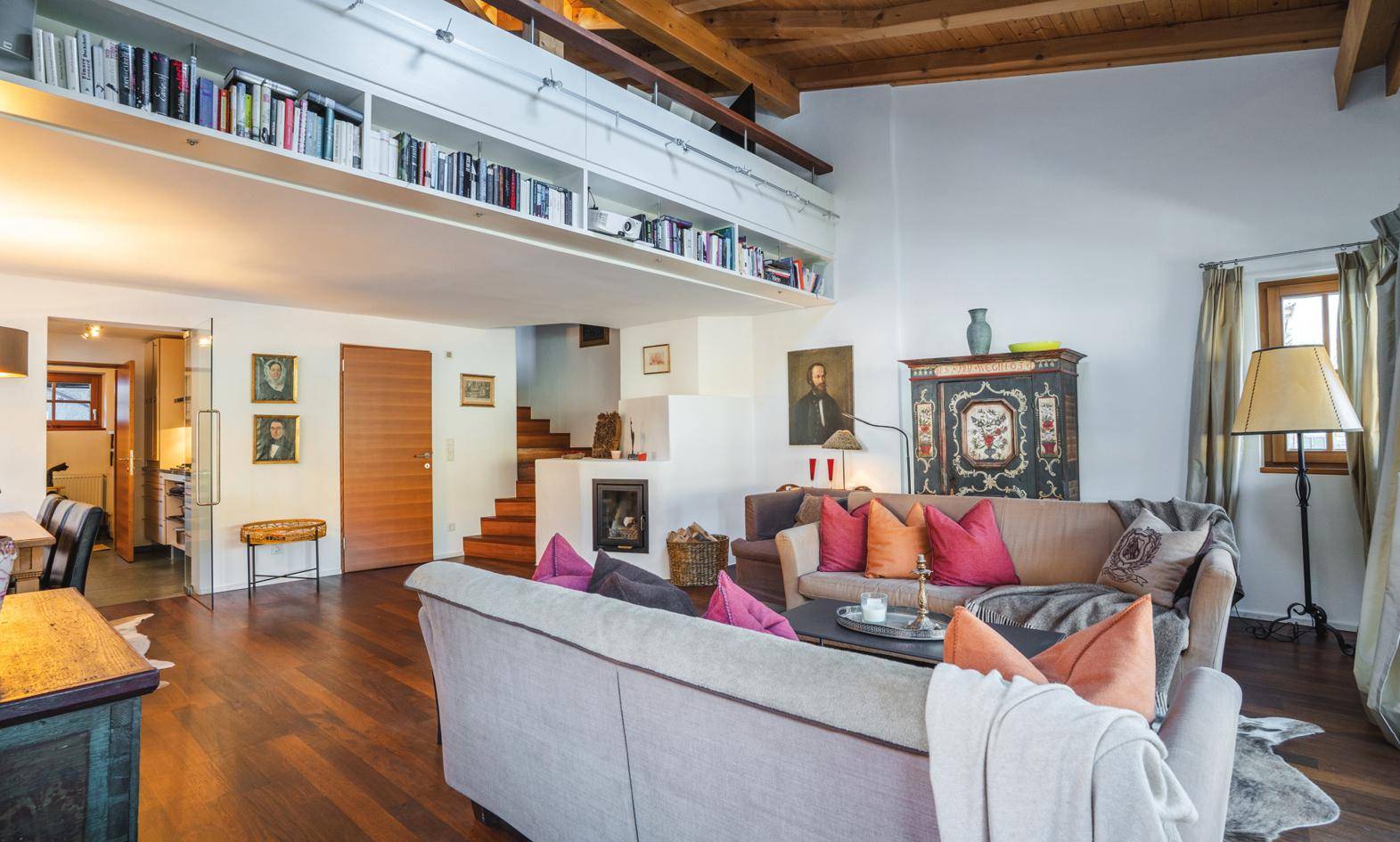 Helles Wohnzimmer mit Holzbalkendecke, Kamin und Zugang zu einer Galerie mit Bücherregalen.