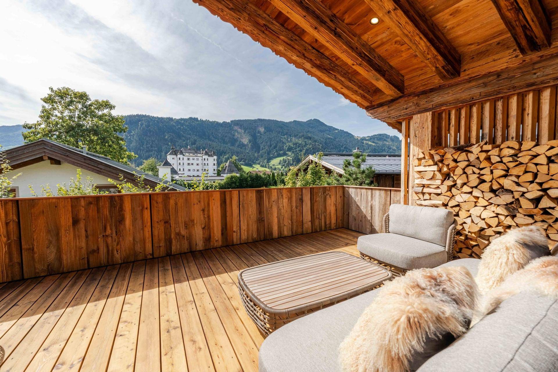 Sonnige Holzterrasse mit bequemen Loungemöbeln und Blick auf die Berge und ein historisches Gebäude.