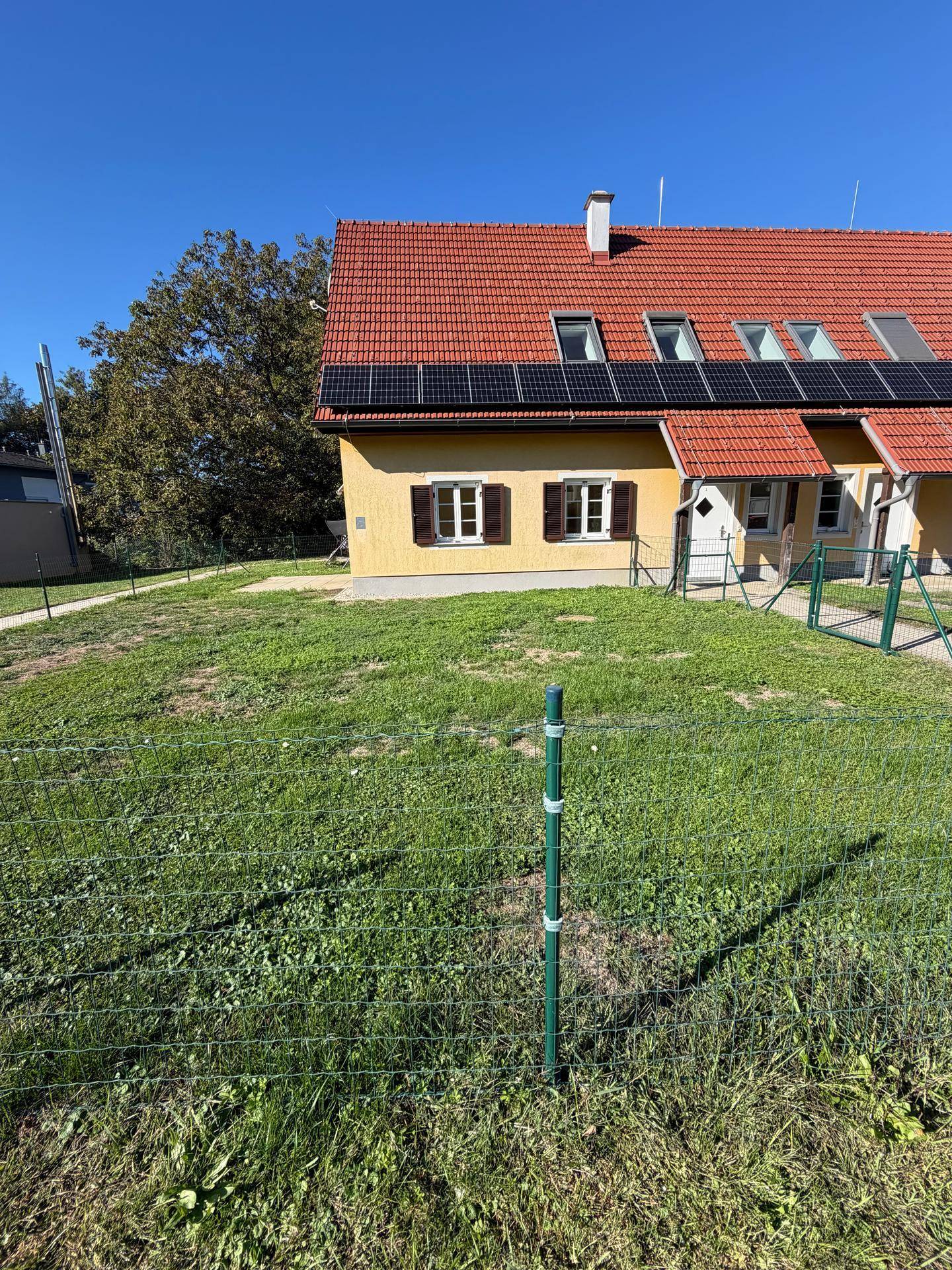 Nahaufnahme der Hausfassade mit Solarpaneelen, Fenstern und einem gepflegten Rasenbereich bei Sonnenschein.