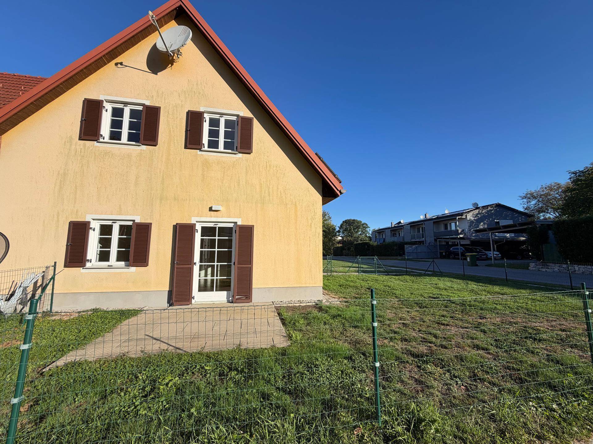 Seitenansicht des Hauses mit gelber Fassade, braunen Fensterläden und Rasenfläche an einem sonnigen Tag.