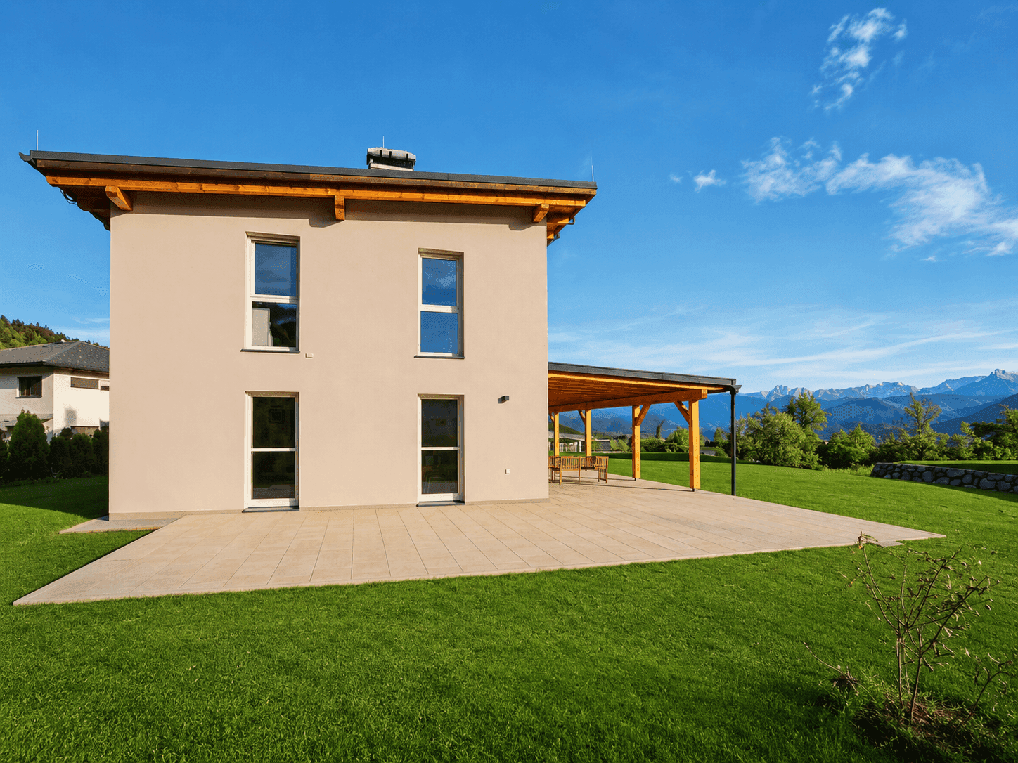 Rückseite des Hauses mit großer, gepflasterter Terrasse und überdachtem Bereich, umgeben von Rasen.