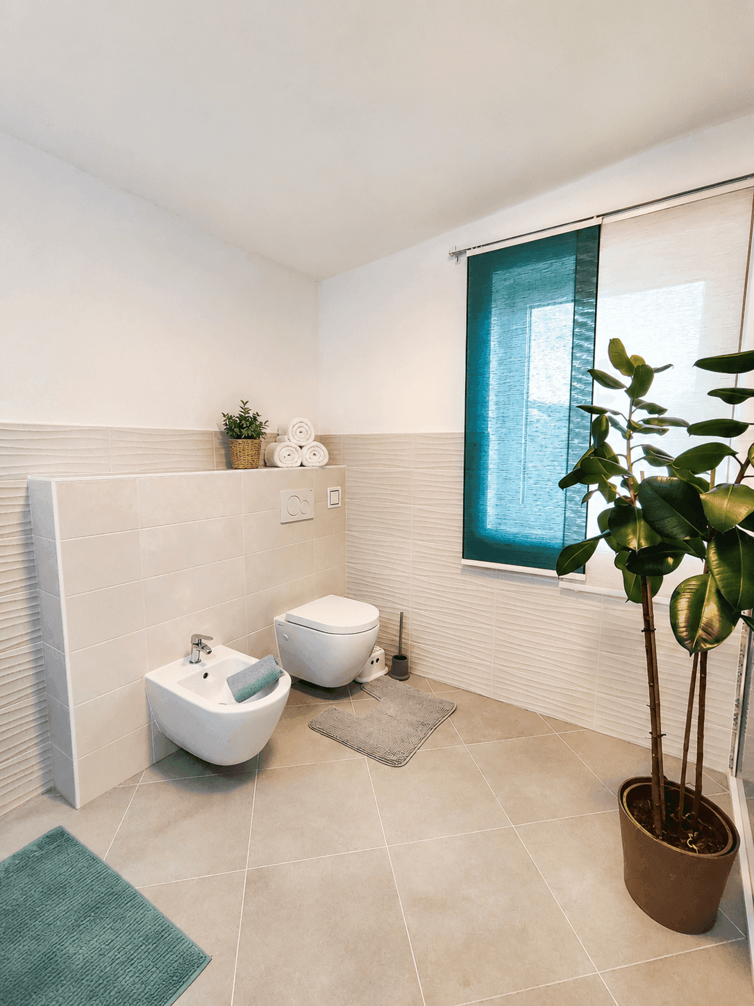 Modernes Badezimmer mit Bidet, Toilette, Fenster und einer großen Grünpflanze, hell gefliest.