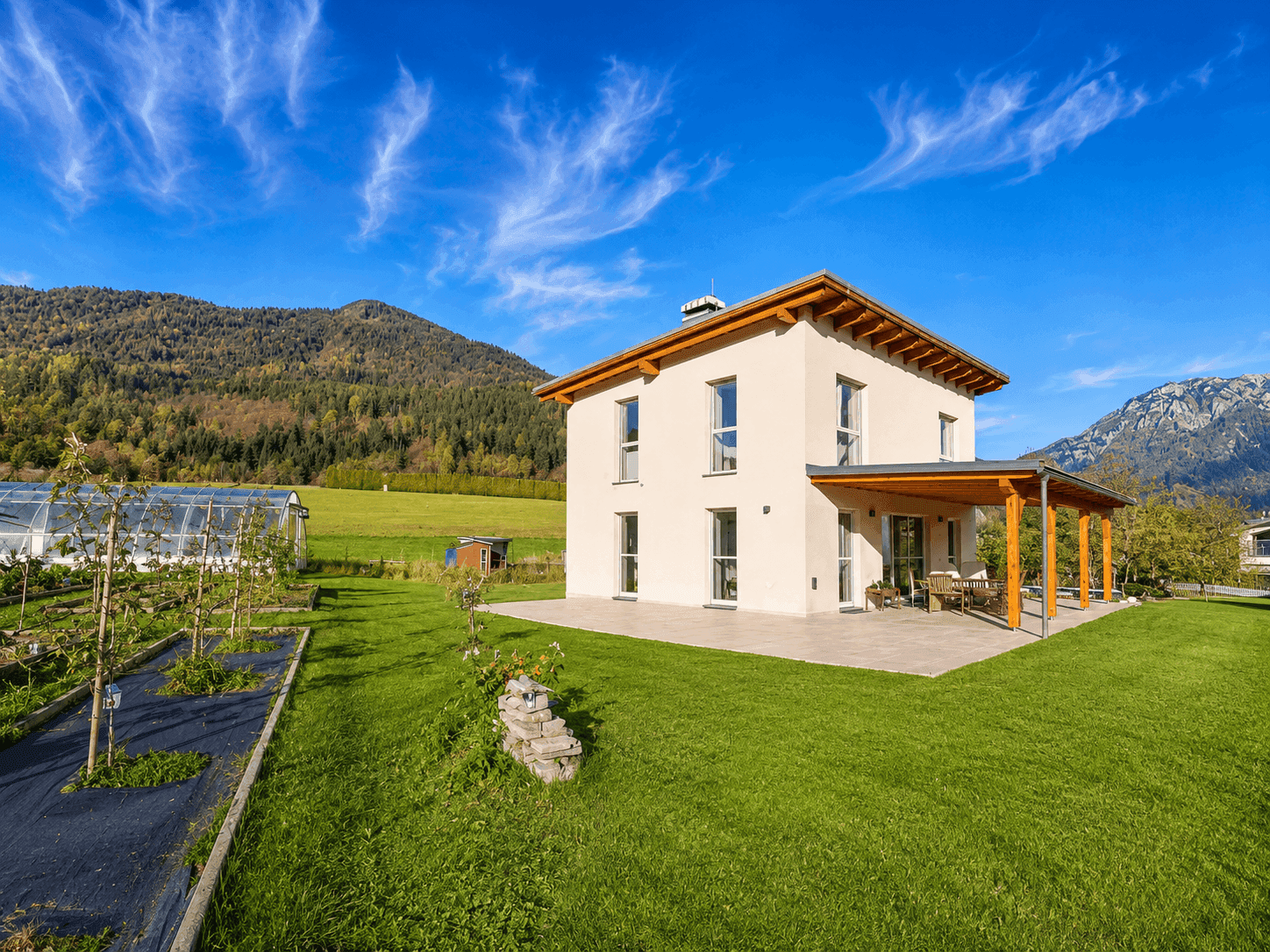 Freistehendes Haus mit großer Rasenfläche, Gewächshaus und überdachter Terrasse vor Bergkulisse.