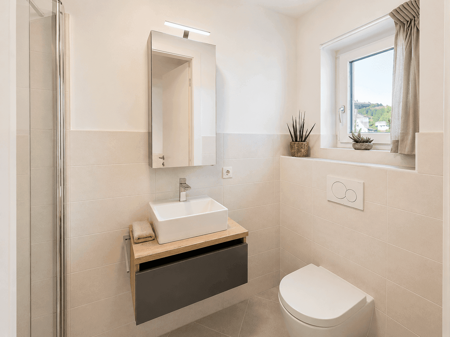 Helles Badezimmer mit modernem Waschtisch, Spiegelschrank, Toilette und Fenster, hell gefliest.
