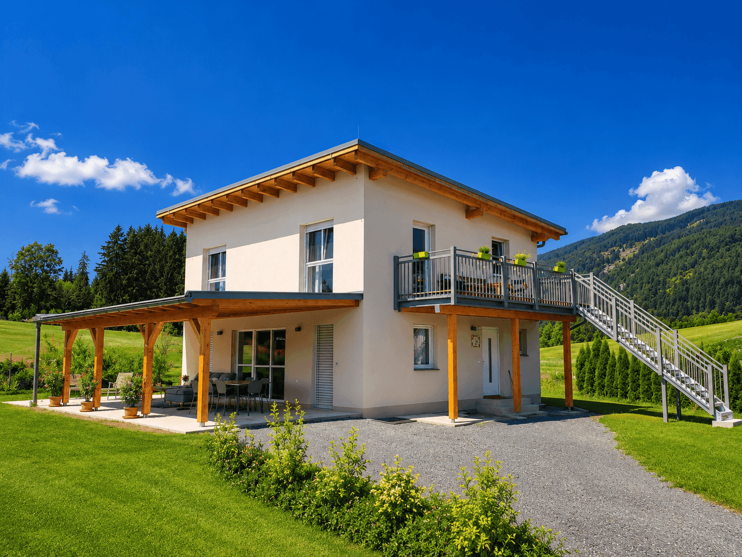 Modernes Wohnhaus mit Balkon und überdachter Terrasse, umgeben von grüner Landschaft und Bergen.