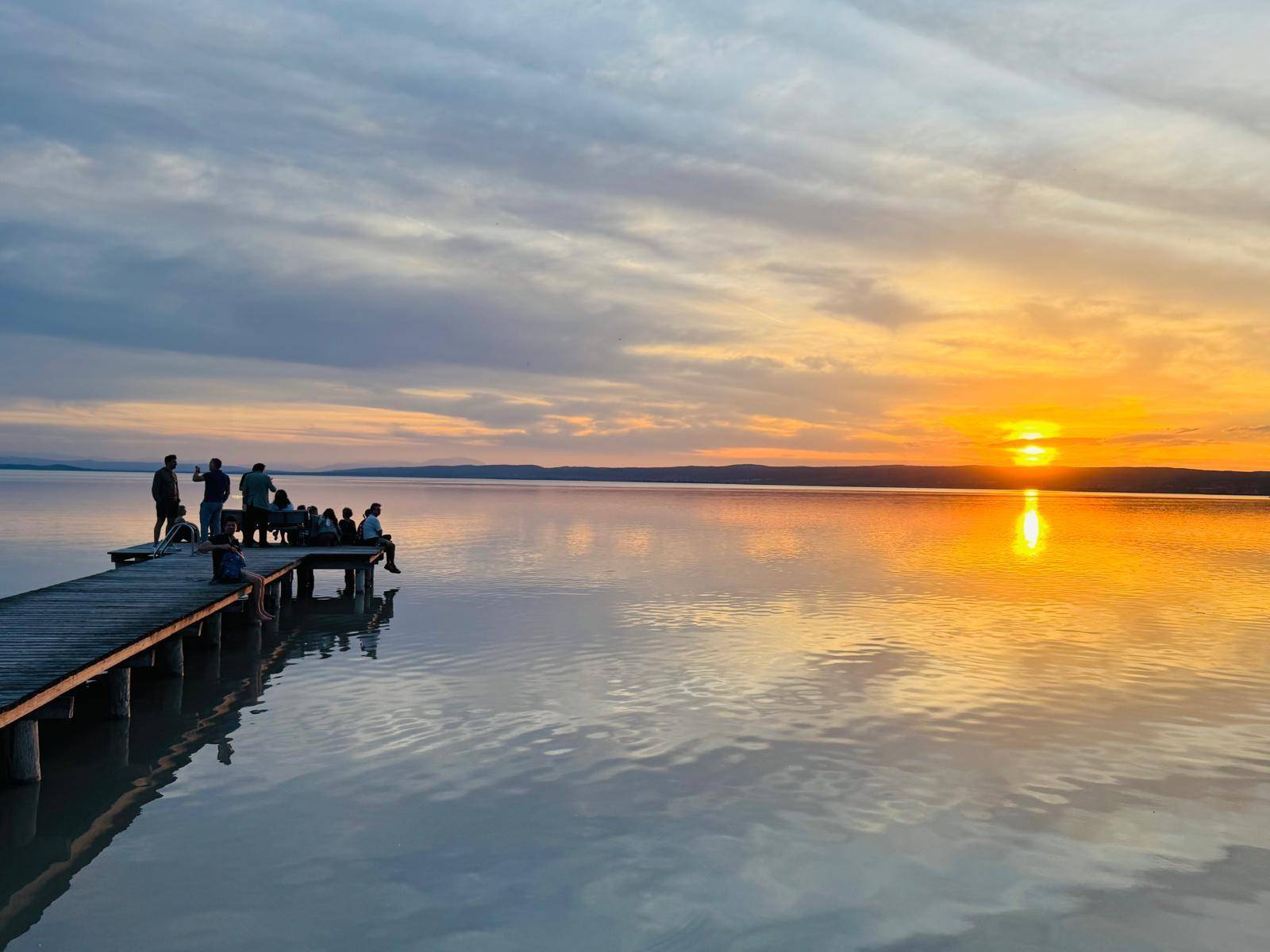 Spektakulärer Sonnenuntergang über einem ruhigen See mit einem Holzsteg und Menschen am Ufer.