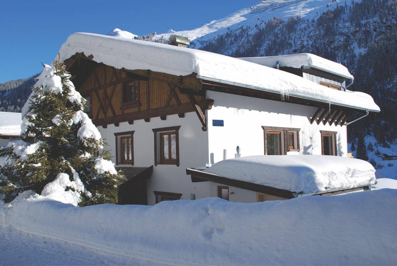 Alpines Chalet mit schneebedecktem Dach und Fassade, umgeben von Winterlandschaft und Bergen.
