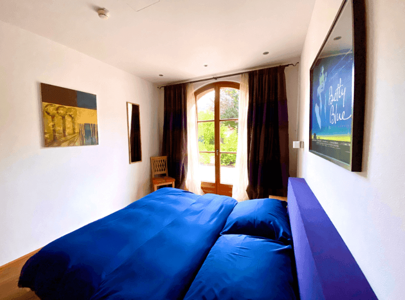 Gemütliches Schlafzimmer mit Holzboden, blauem Bett und großen Fenstern, die viel Licht hereinlassen.