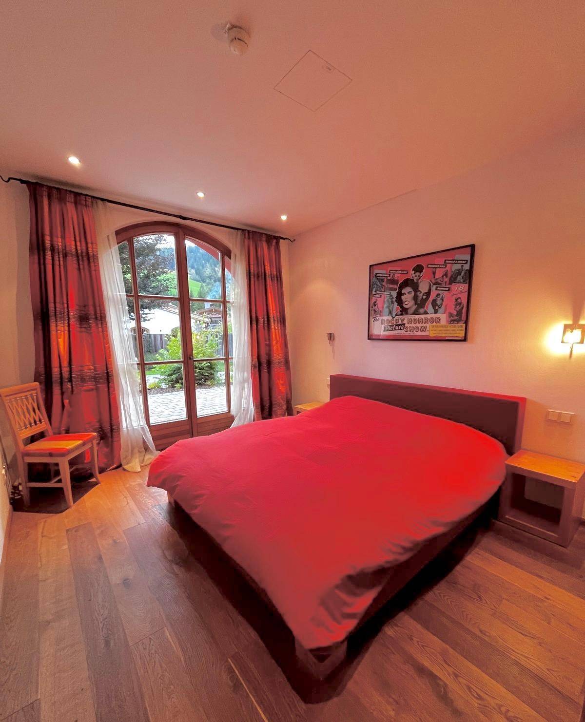 Schlafzimmer mit rotem Bett, Holzboden und großen Fenstern mit Zugang zum Garten.