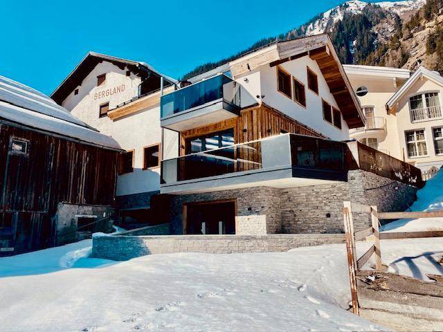 Rückansicht eines modernen Gebäudes mit Glasbalkonen und Steinfundament, eingebettet in eine verschneite Berglandschaft.