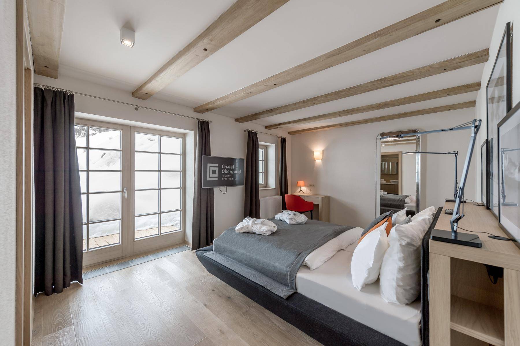 Helles Schlafzimmer mit Holzbalken, großem Bett und Fenstertür zum verschneiten Außenbereich.