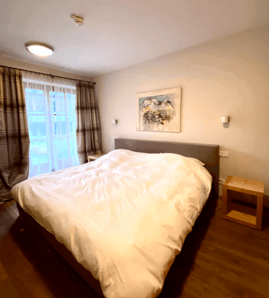 Schlafzimmer mit Doppelbett, Holzboden und Vorhängen vor dem Fenster, das Tageslicht spendet.