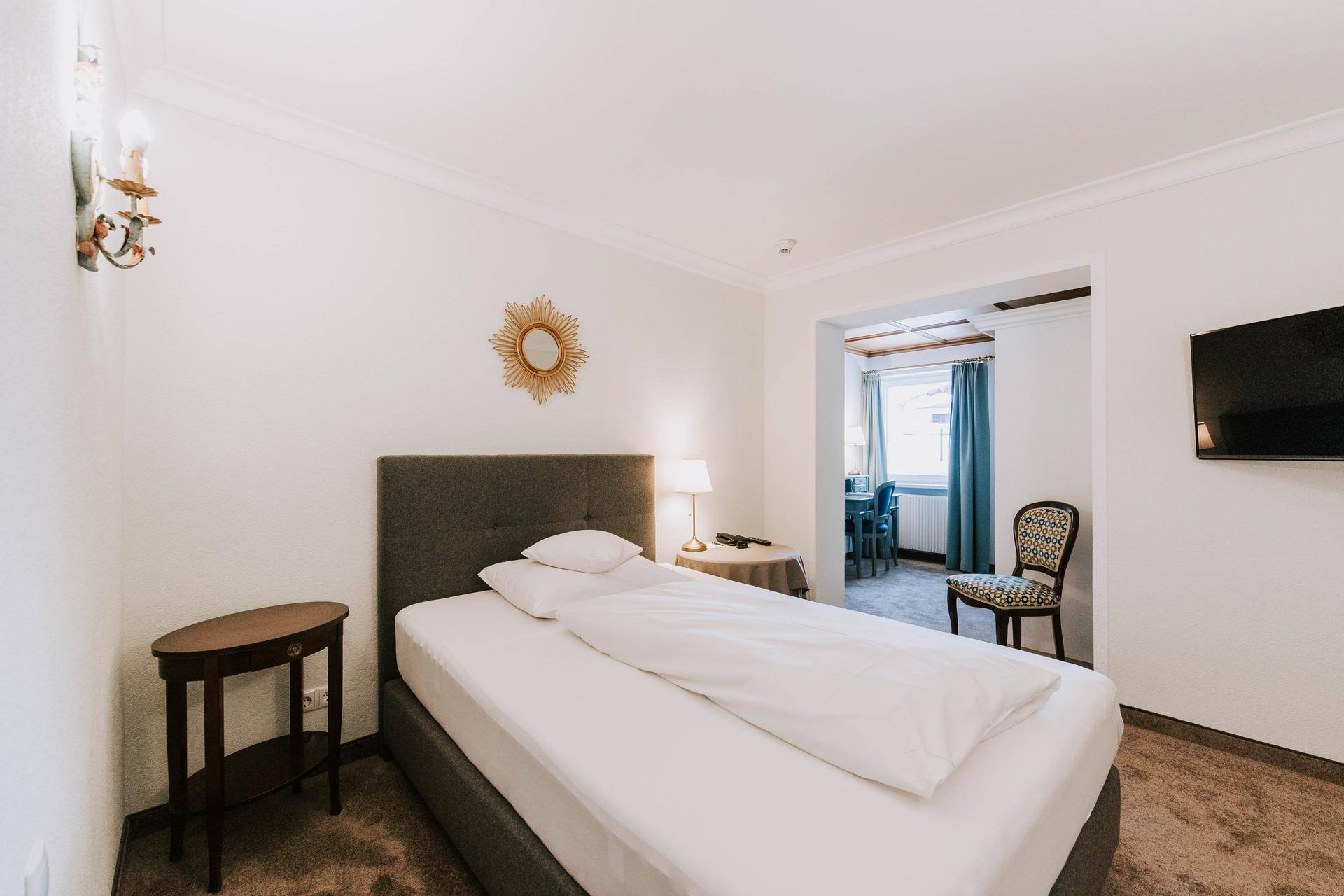 Einzelzimmer mit Bett, Nachttisch und Teppichboden, angrenzend an einen weiteren Raum.