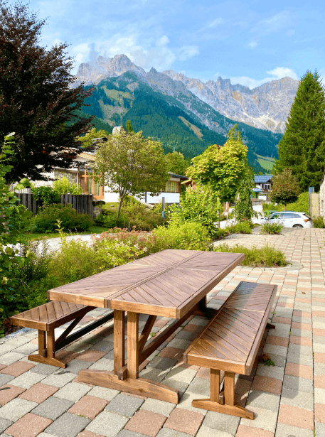 Sonniger Außenbereich mit Holztisch und Bänken, umgeben von grüner Natur und Bergblick.