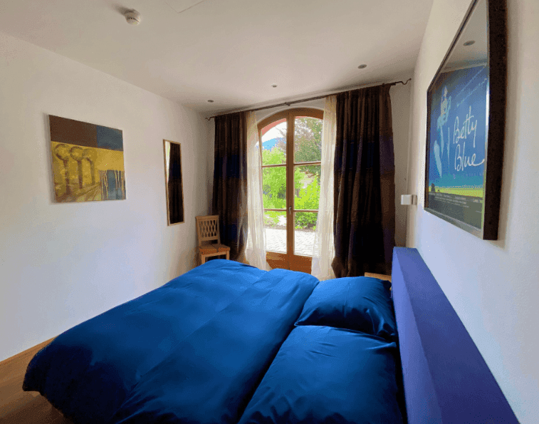 Schlafzimmer mit Holzboden, blauem Bett und großen Fenstern mit Blick ins Grüne.