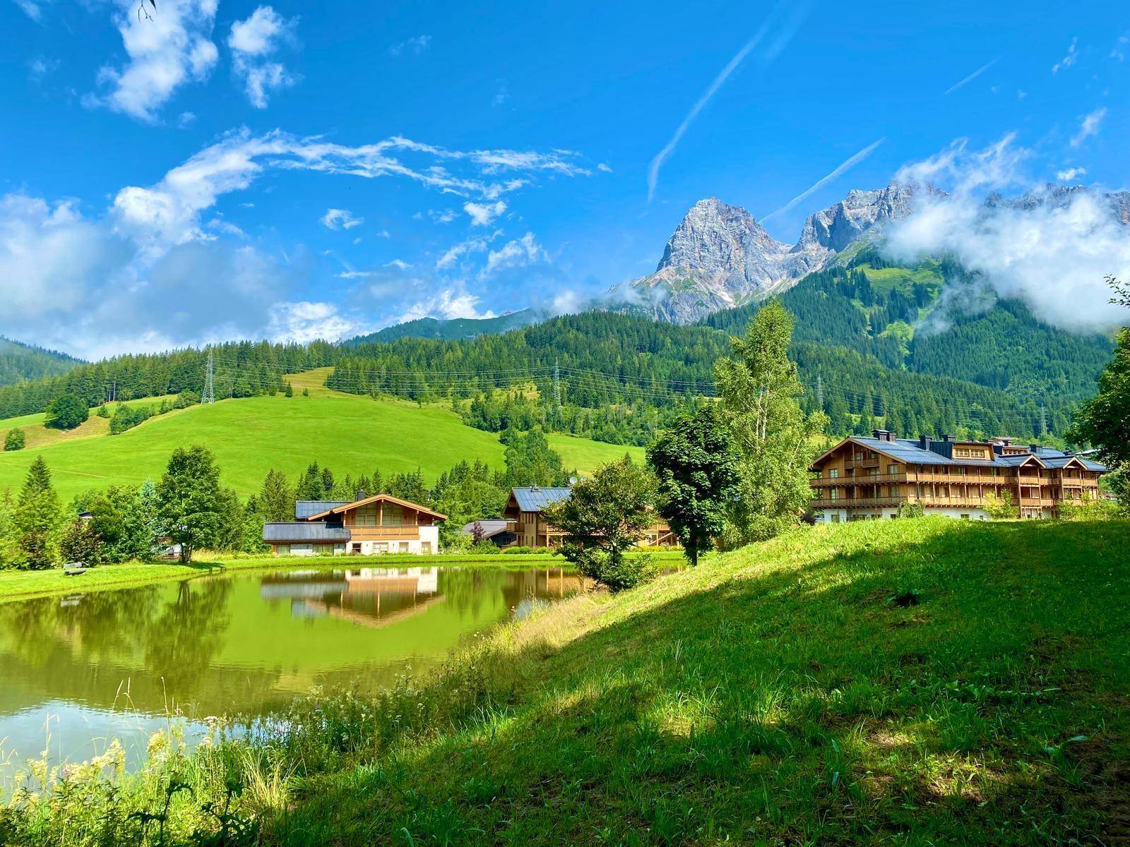 Malerische Landschaft mit See, grünen Hügeln und traditionellen Häusern vor einer Bergkulisse.
