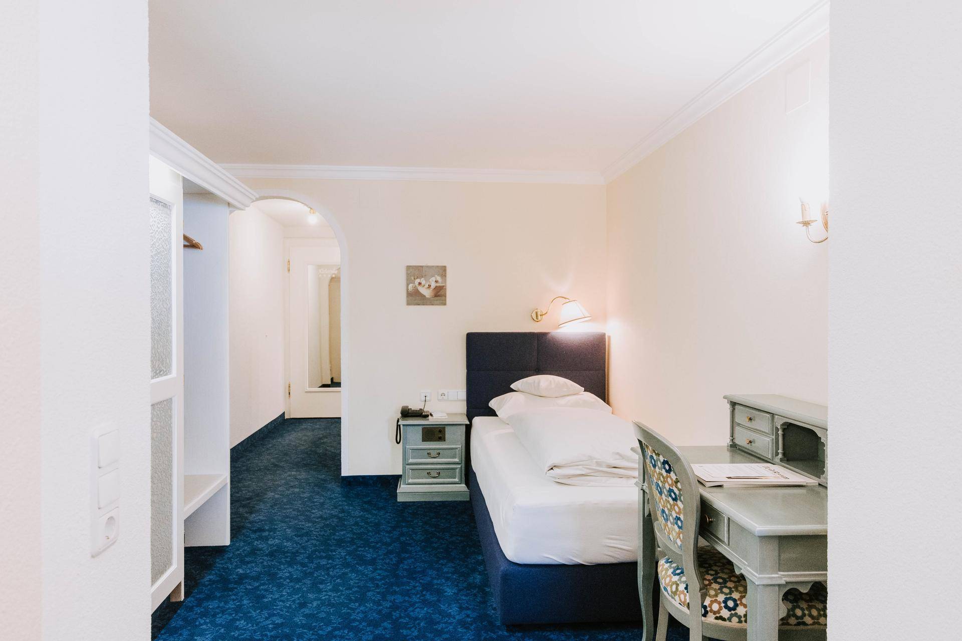Einzelzimmer mit Bett, Schreibtisch und blauem Teppichboden, angrenzend an einen Flur.