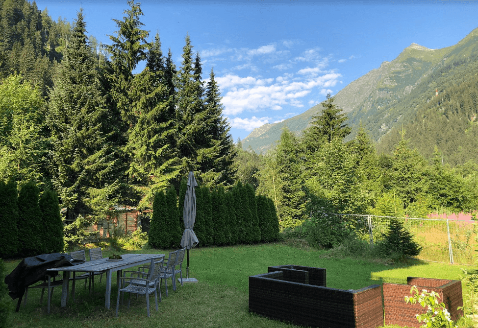 Grüner Garten mit Sitzgelegenheiten und Bergblick, umgeben von hohen Bäumen.