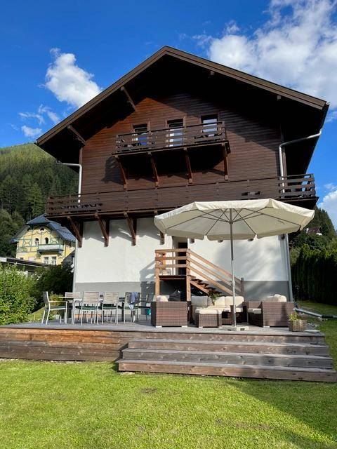 Große Holzterrasse mit Gartenmöbeln und Sonnenschirm, angrenzend an einen grünen Rasen.