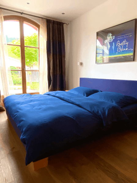 Helles Schlafzimmer mit blauem Bett, Holzboden und Zugang zum Garten durch große Fenster.