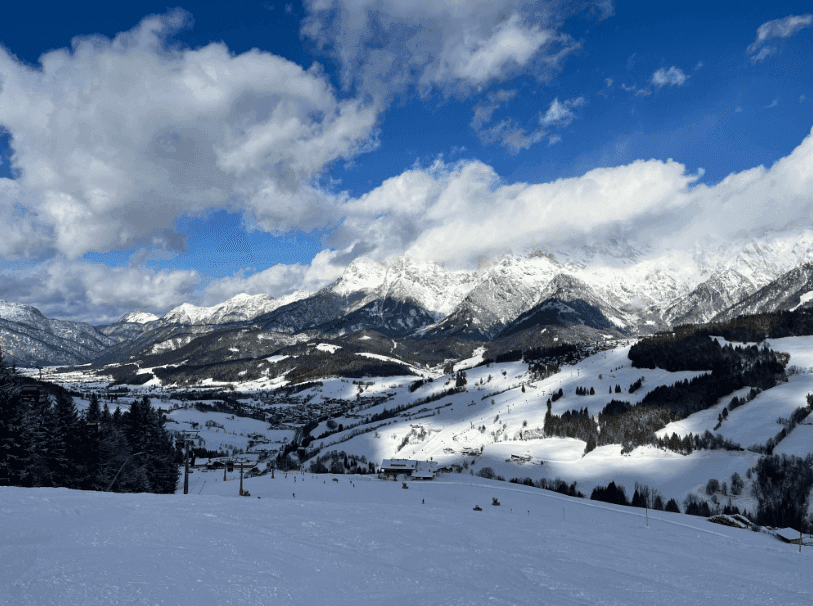 Weitläufige, verschneite Berglandschaft mit Skipisten und bewaldeten Hängen unter blauem Himmel.