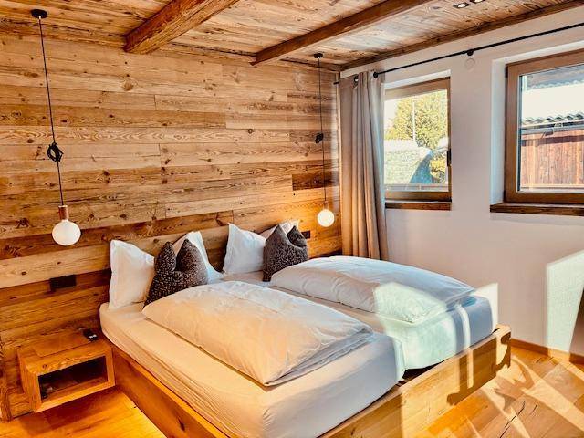 Helles Schlafzimmer mit Holzwänden, Holzbalkendecke und zwei Fenstern, die viel Tageslicht hereinlassen.