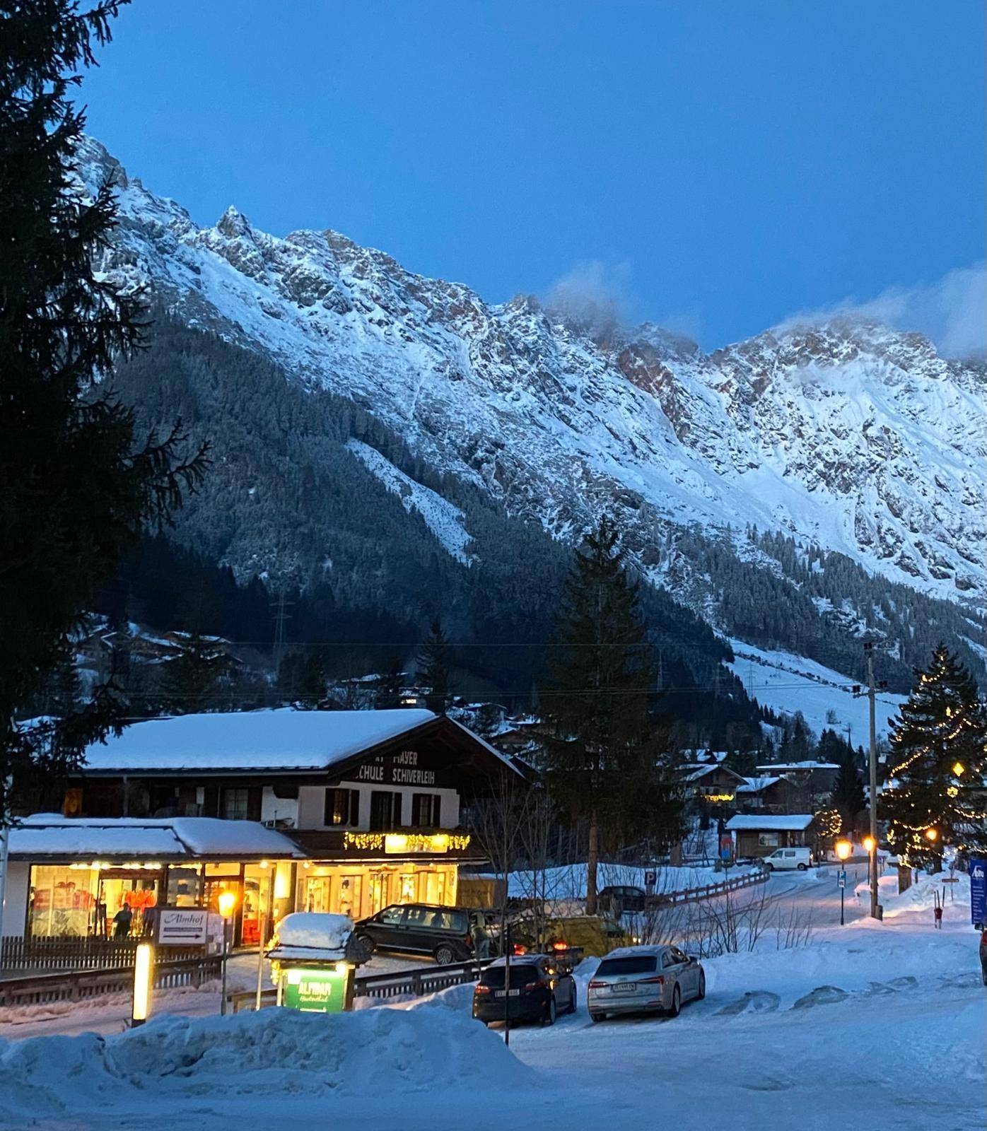 Verschneite Dorfstraße bei Dämmerung mit beleuchteten Gebäuden und majestätischen Bergen im Hintergrund.