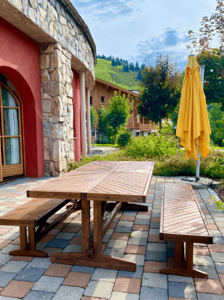 Gemütliche Terrasse mit Holztisch und Bänken, umgeben von grüner Natur und Bergblick.