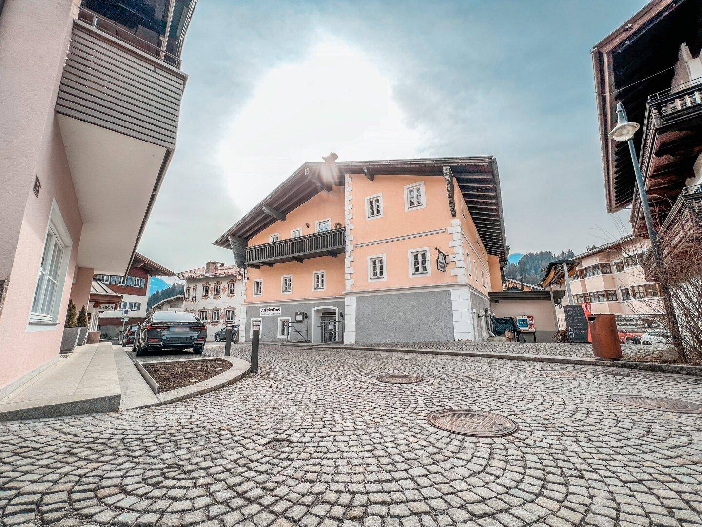 Weitwinkelansicht eines traditionellen Gebäudes mit Balkon und gepflasterter Straße in einer bergigen Umgebung.