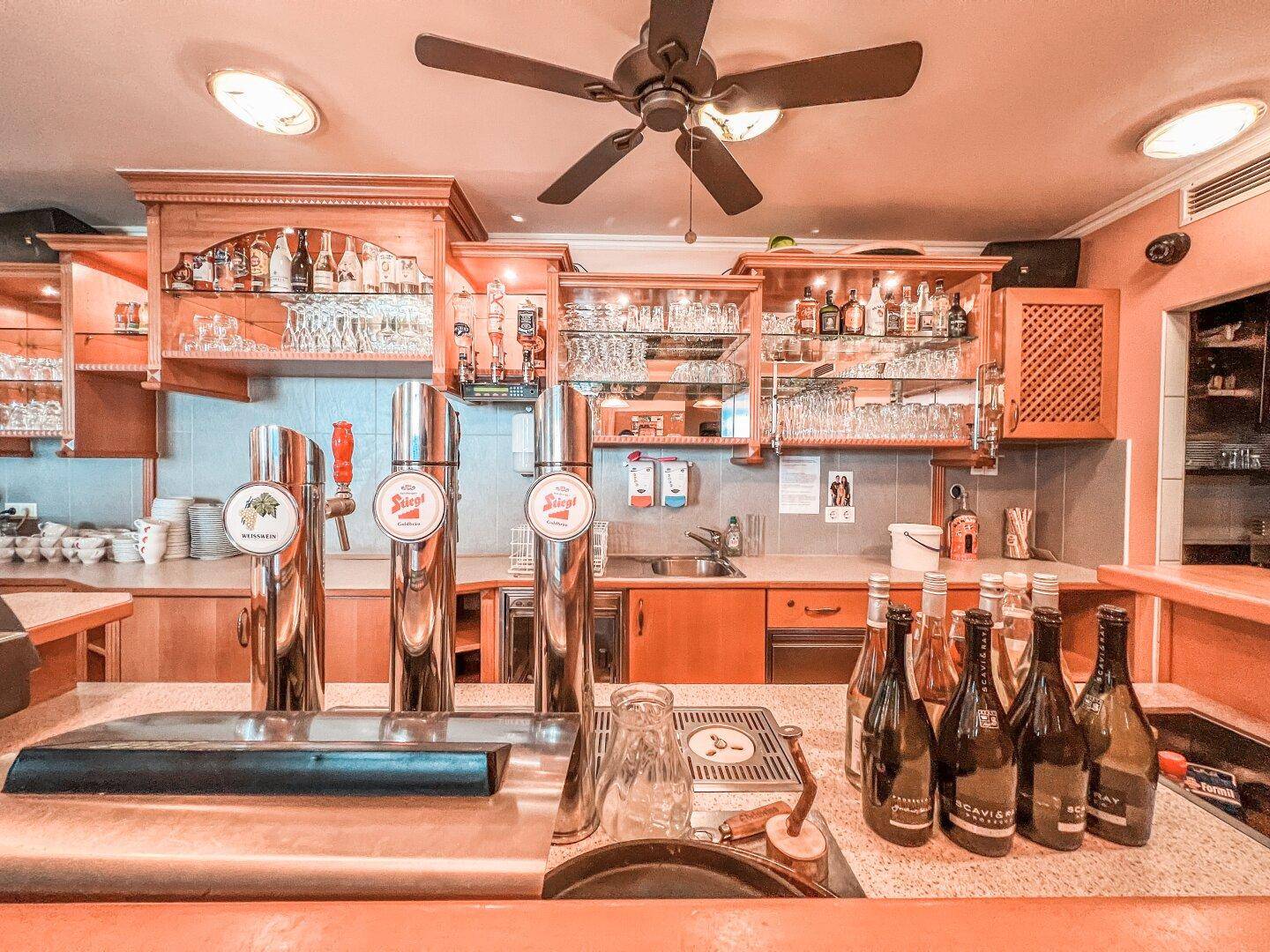Detailansicht des Barbereichs mit glänzenden Zapfanlagen, Gläsern und Flaschen unter einem Deckenventilator.