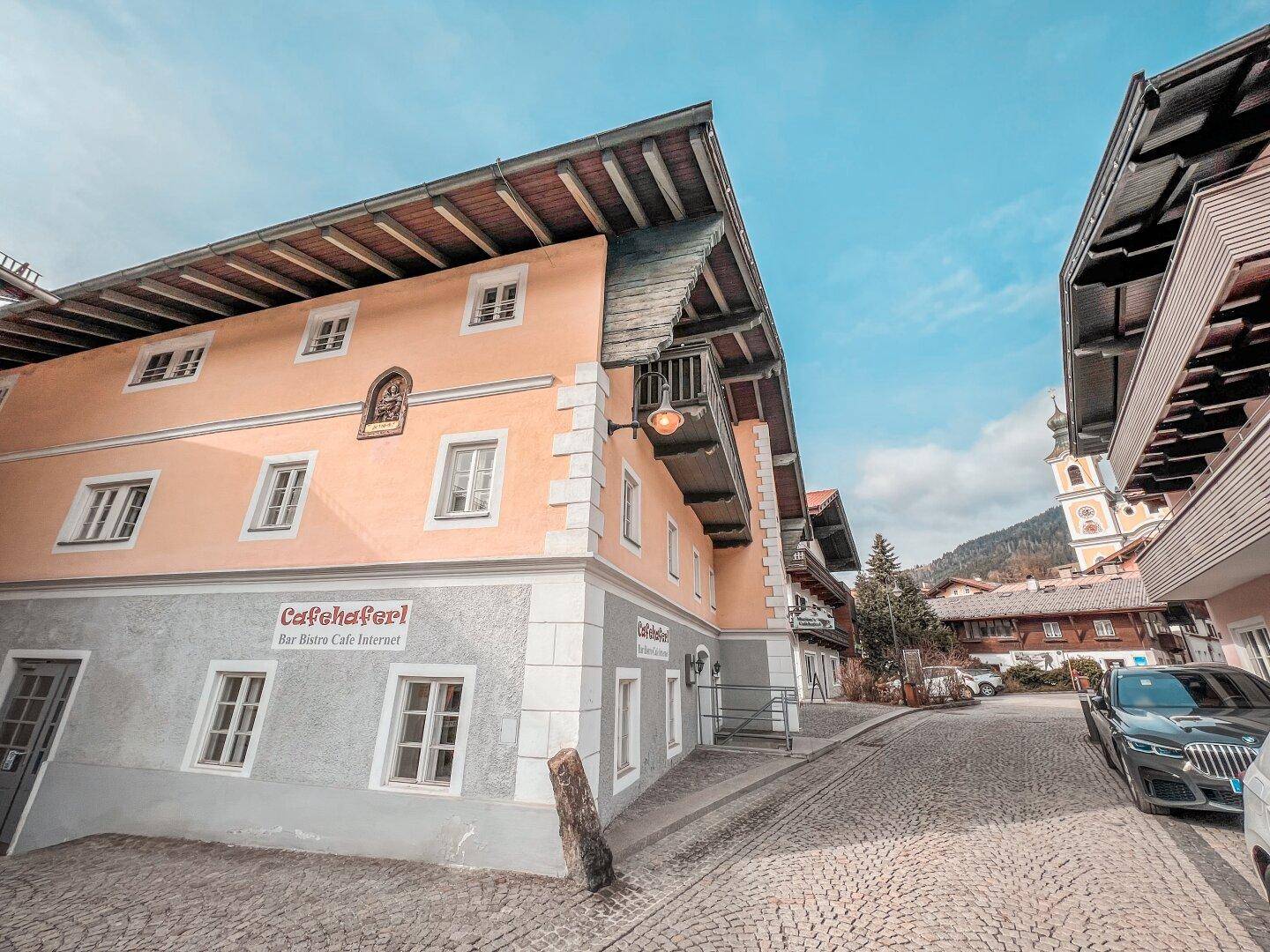Traditionelles Gebäude mit Balkon und hellrosa Fassade an einer gepflasterten Straße, mit Kirchturm im Hintergrund.