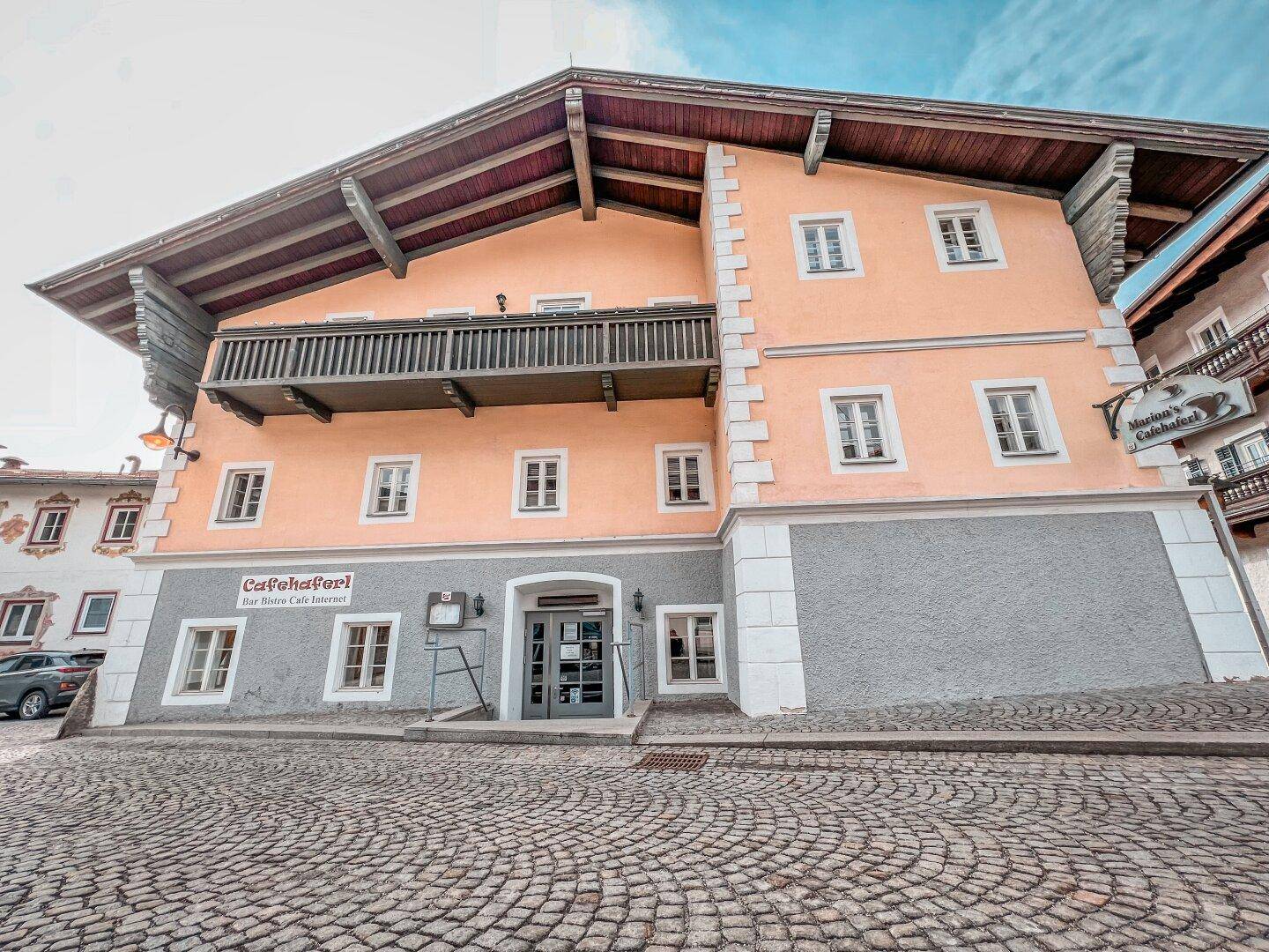 Historisches Gebäude mit Holzbalkon und gepflastertem Vorplatz, umgeben von traditioneller Architektur.