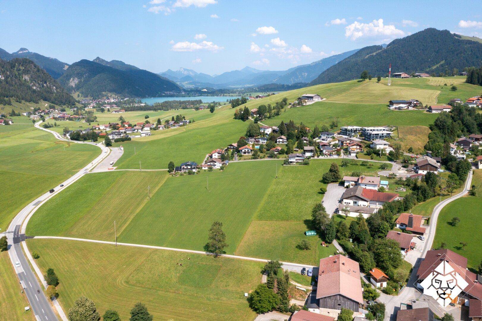 Weitläufige Luftaufnahme eines Dorfes in einem Tal, umgeben von grünen Feldern, Bergen und einem See.