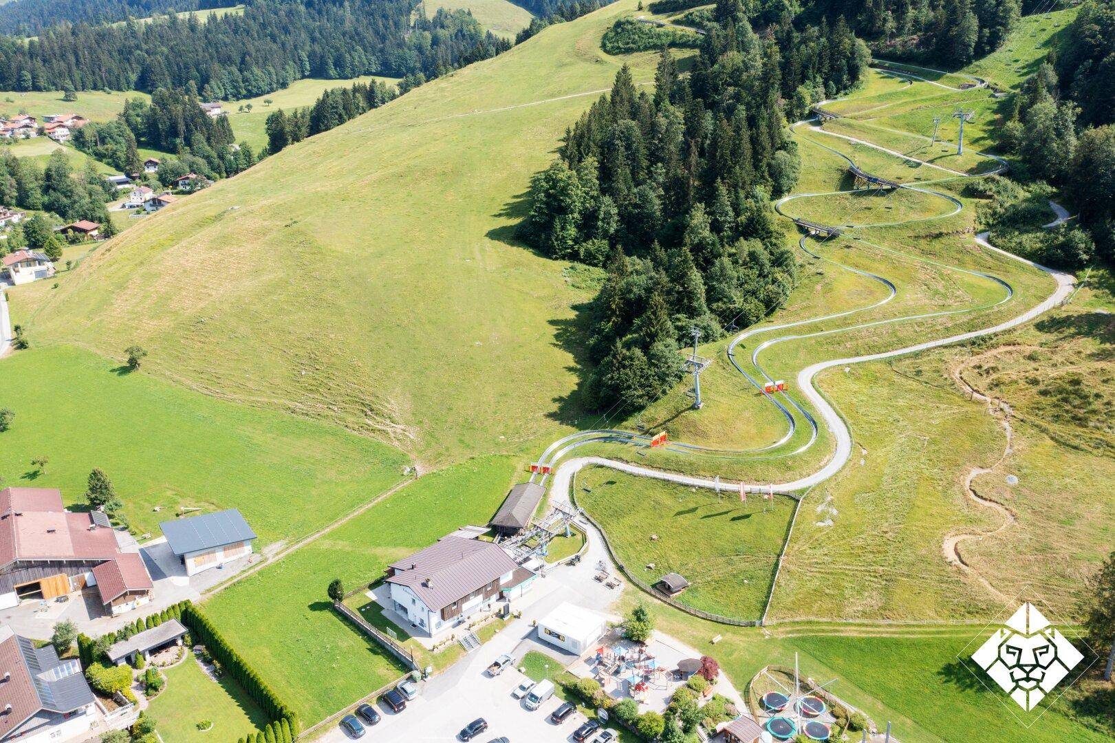 Luftaufnahme einer Sommerrodelbahn, die sich durch eine grüne Hügellandschaft schlängelt, neben Gebäuden und Parkplätzen.