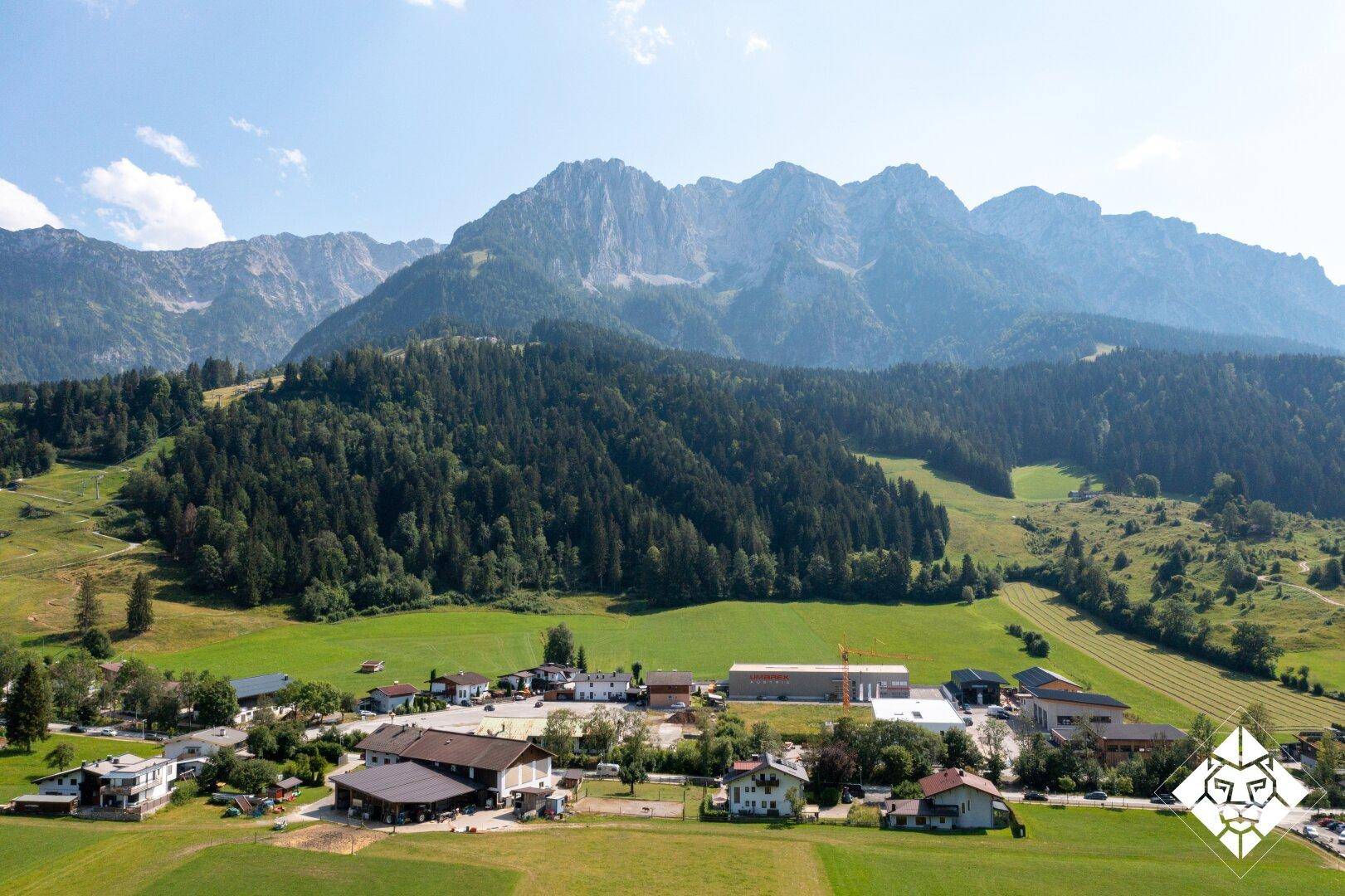 Luftaufnahme einer ländlichen Siedlung mit beeindruckender Bergkulisse, umgeben von grünen Wiesen und Wäldern.