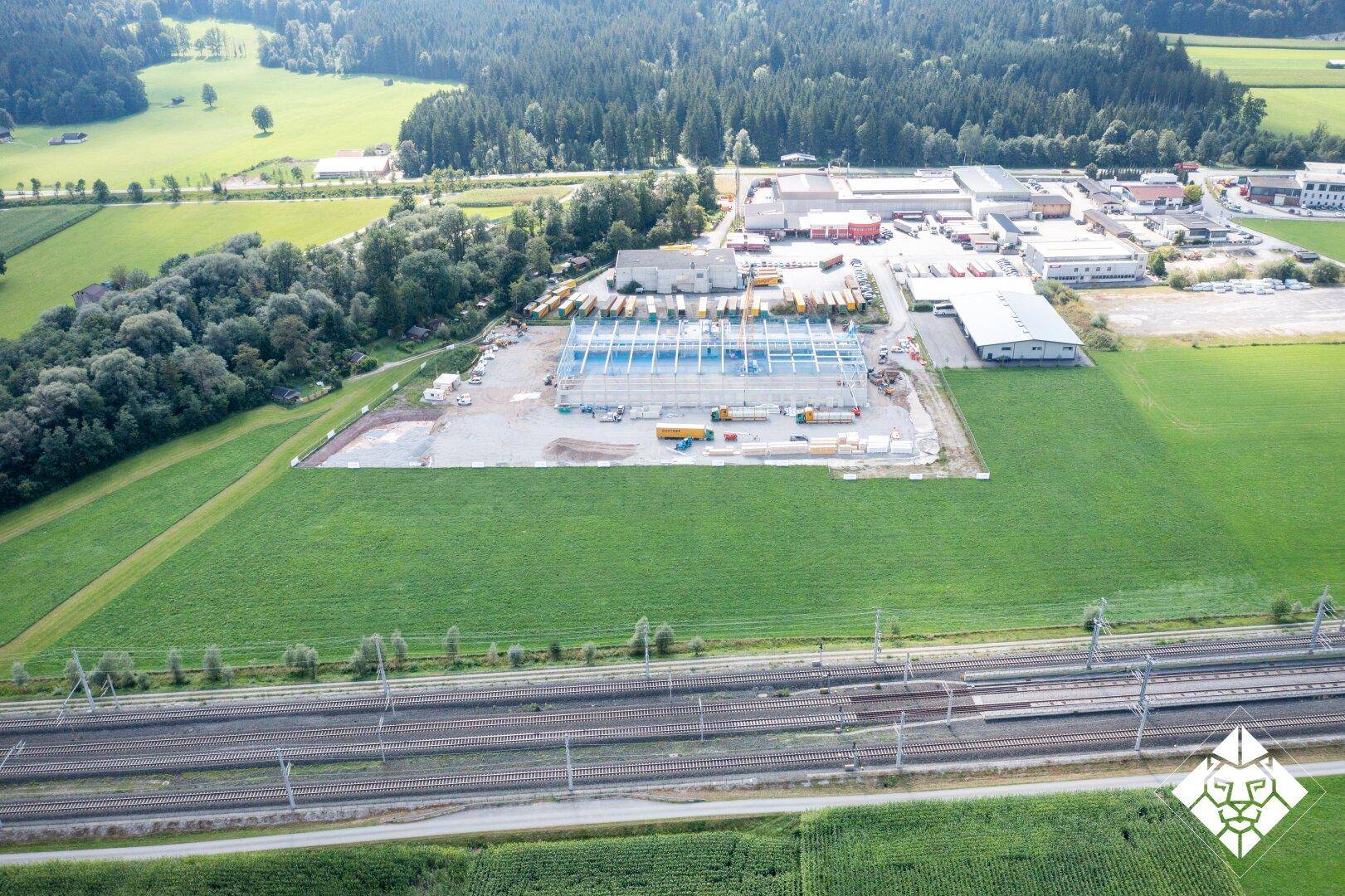 Panoramablick aus der Luft auf eine Baustelle und angrenzende Industriegebäude, umgeben von Feldern und Bahngleisen.