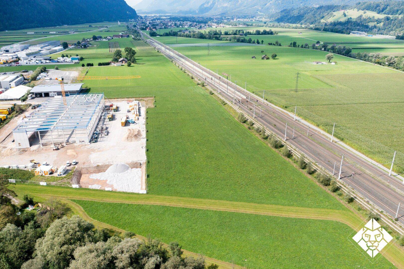 Weitläufige Luftaufnahme einer Baustelle neben Bahngleisen, eingebettet in eine ländliche Landschaft mit Bergen.