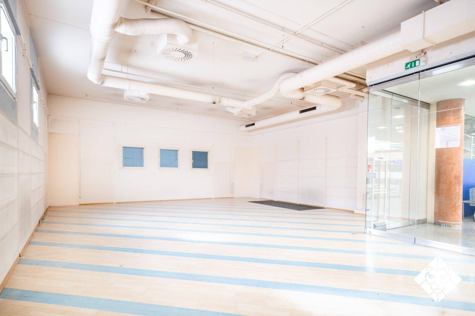 Leerer, heller Gewerberaum mit freiliegenden Rohren an der Decke, kleinen Fenstern und gestreiftem Holzboden.
