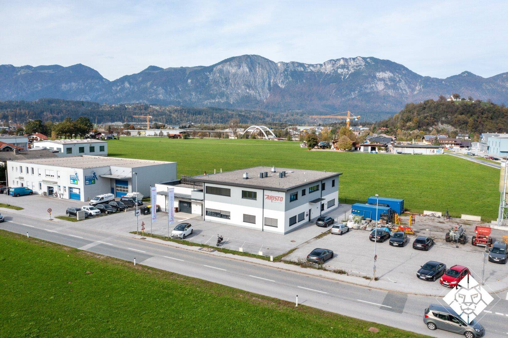 Luftaufnahme eines Gewerbegebiets mit mehreren Gebäuden, Parkplätzen und Bergen im Hintergrund.