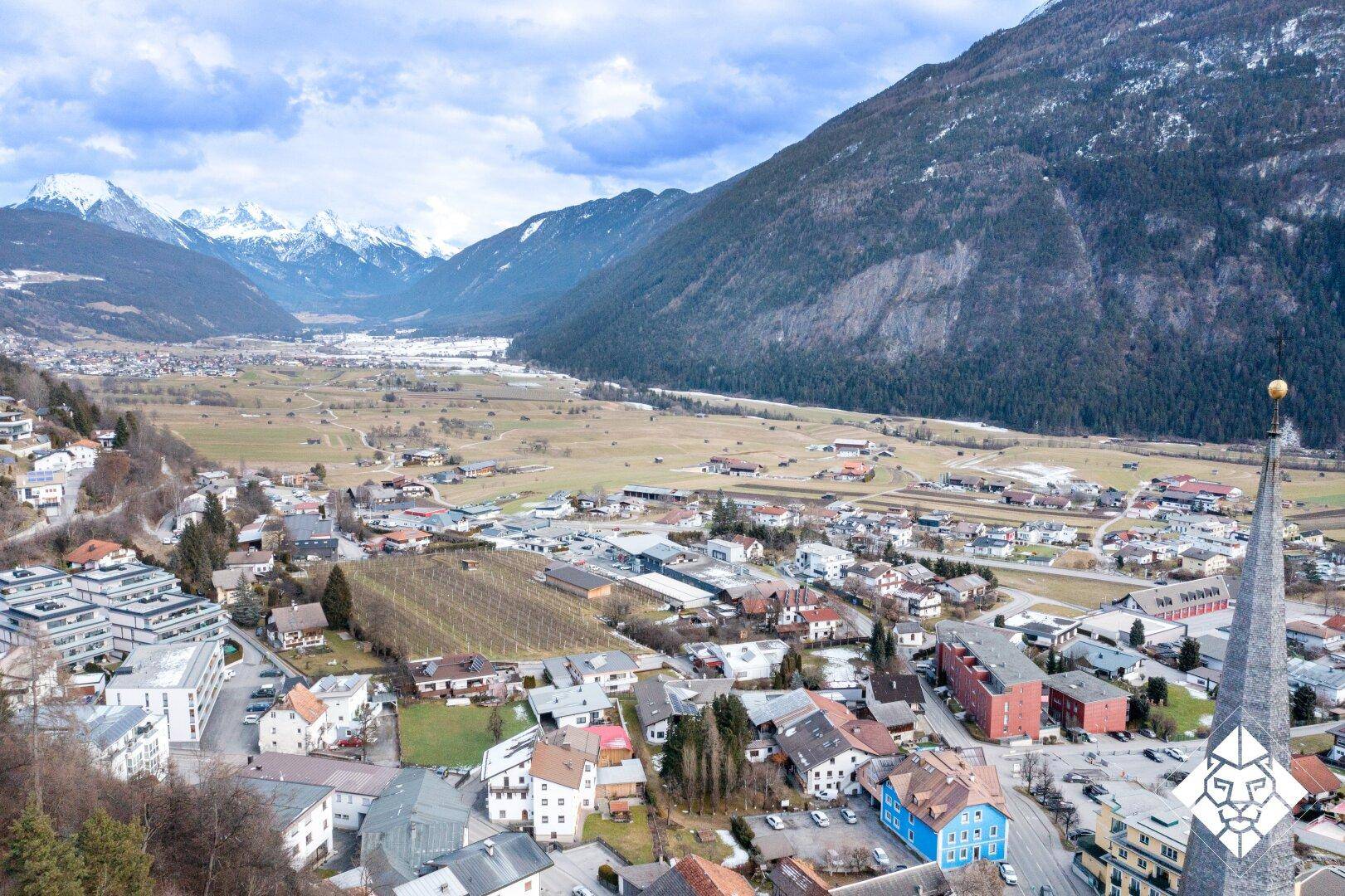 Weitwinkel-Luftaufnahme einer Stadt in einem Tal, umgeben von bewaldeten Hügeln und schneebedeckten Bergen.