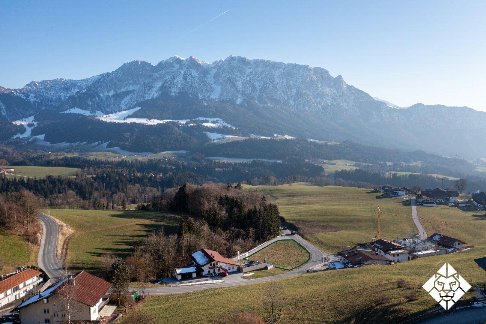 Panoramablick auf die weitläufige Landschaft mit schneebedeckten Bergen, grünen Feldern und dem markierten Baugrundstück.