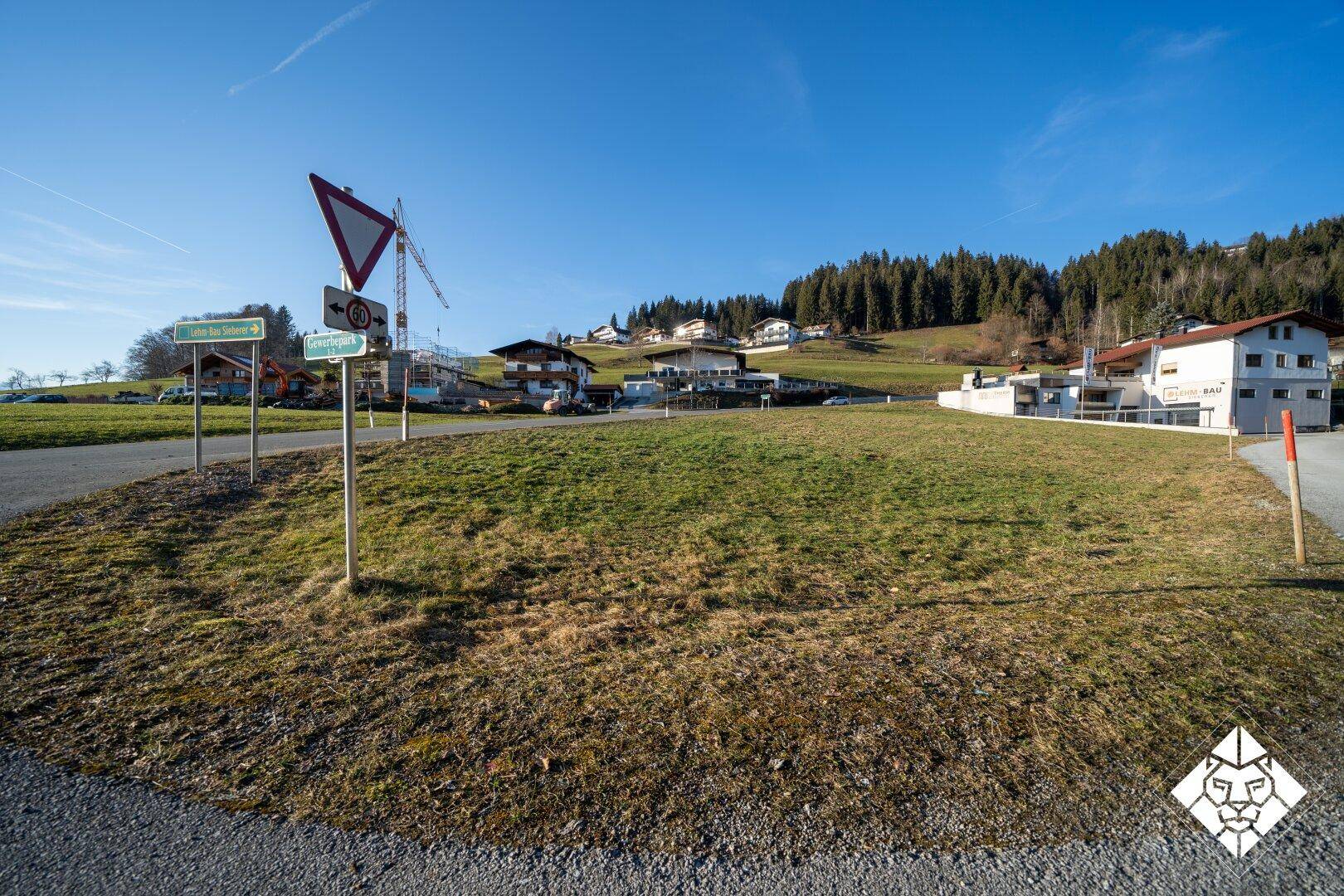 Blick auf das grüne Baugrundstück von der Straße aus, umgeben von Wohnhäusern und einer hügeligen Landschaft.