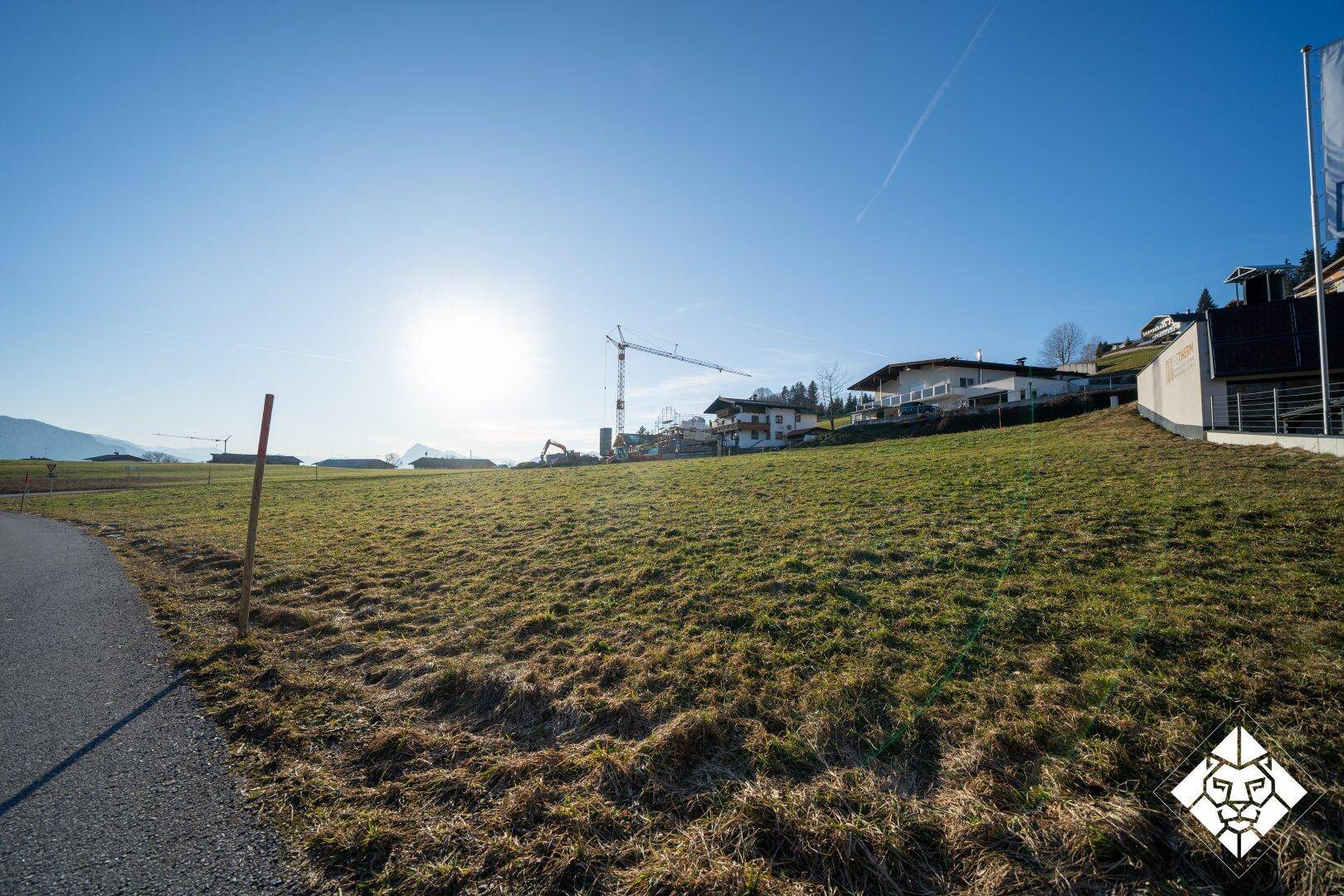 Weitläufiges, sonniges Baugrundstück mit Blick auf eine Baustelle und Häuser am Hang in der Ferne.