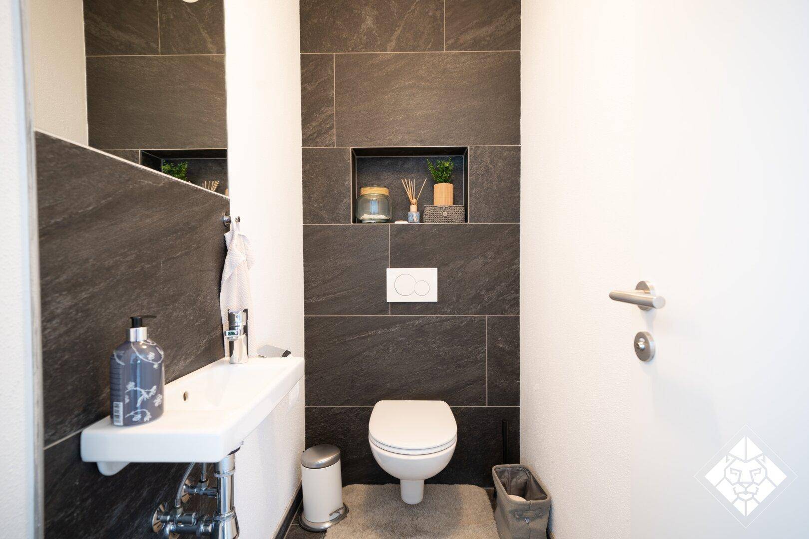 Kleine Gästetoilette mit Wand-WC, Handwaschbecken und dunklen großformatigen Fliesen an der Wand.