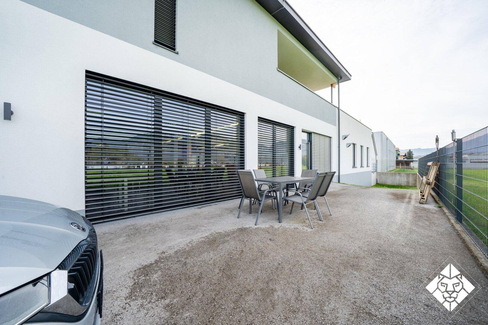 Außenbereich mit Terrassenmöbeln und großen Fenstern, umgeben von einem Zaun.